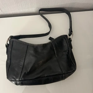 Svart axelväska i skinn - Snygg svart axelväska i äkta skinn med enkel design och silverfärgade metalldetaljer. Väskan har dragkedja upptill och ett innerfack, perfekt storlek för det viktigaste. Stilren och passar till många outfits.