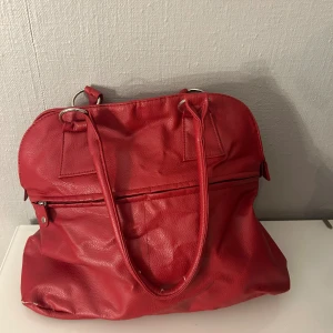 Röd axelväska i skinnlook - Cool röd axelväska med långa handtag och silverfärgade ringdetaljer. Väskan har en dragkedja framtill och ett stort huvudfack med innerficka. Perfekt för dig som vill ha plats för allt du behöver och samtidigt sticka ut med färg.