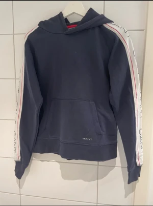 Marinblå hoodie från GANT med logga - Snygg marinblå hoodie från GANT med huva och klassisk känguruficka. Längs ärmarna finns vita band med röd linje och GANT-logga. Perfekt för dig som gillar sportig och avslappnad stil. Mjuk bomullskvalitet och långärmad modell.