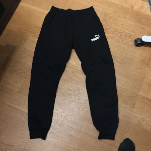 Svarta mjukisbyxor från Puma, stl S - Svarta mjukisbyxor från Puma med logga i vitt på vänster lår. Byxorna har ribbade muddar vid bensluten och elastisk midja för skön passform. Perfekta för chill eller träning, tillverkade i mjukt bomullsmaterial.