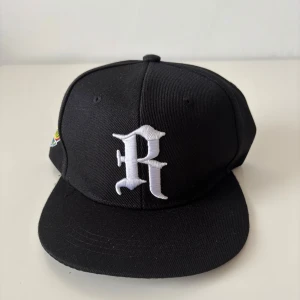 New Era liknande keps - Svart snapback keps med platt skärm och broderat vitt R på framsidan. Justerbar baktill med plastspänne och färgglad broderad detalj på sidan. Snygg och stilren design som passar till streetwear. Materialet är troligtvis bomull eller polyester. Helt ny.