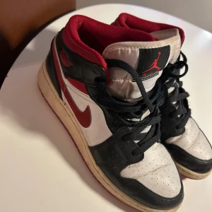 Nike Air Jordan 1 High röd/svart/vit - Säljer ett par Nike Air Jordan 1 High sneakers i klassisk röd, svart och vit färg. Skorna har snörning, rund tå och platt sula. Ovandelen är i skinn med mesh vid plösen och insidan. Ikonisk Jumpman-logga på plösen och Air Jordan-märke vid sidan.