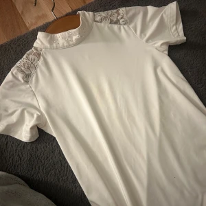 Vit kortärmad tävlingströja med spets och pärlor - Vit kortärmad topp med hög krage dekorerad med pärlor och små stenar. Axelpartierna har inslag av blommig spets i vitt. Materialet känns mjukt och stretchigt, perfekt för en stilren look med extra detaljer. Perfekt tävlings tröja men även för träningar och andra aktiviteter.