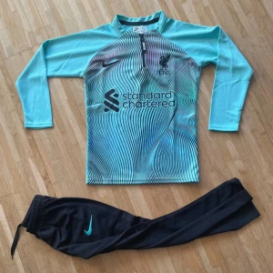 Liverpool långärmad fotbollströja Nike - Säljer en långärmad Liverpool fotbollströja från Nike i ljusblått med coolt vågmönster och svarta detaljer. Tröjan har halv dragkedja fram, Liverpool FC-logga och sponsortryck. Materialet är lätt och andas, perfekt för träning eller match.