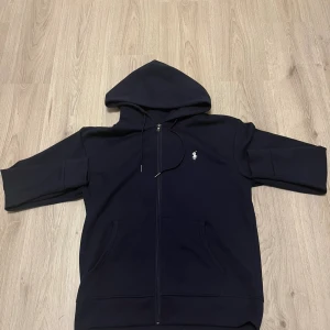 Svart hoodie från Polo Ralph Lauren - Svart hoodie från Polo Ralph Lauren med dragkedja och huva. Klassisk broderad logga på bröstet och kängurufickor framtill. Tillverkad i mjuk bomullsmix, perfekt för chill dagar. Snygg och enkel att matcha med allt.