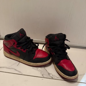 Nike Air Jordan 1 svart/röd sneakers - Nike Air Jordan 1 sneakers i svart och rött med klassisk high-top siluett. Skorna har snörning, rund tå och platt sula. Ikonisk Air Jordan-logga på sidan och röd Swoosh. Tillverkade i skinn med svart textilfoder. Perfekt för dig som gillar streetwear och basketstil.
