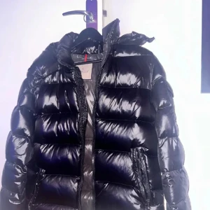  Moncler maya pufferjacka - Säljer en svart pufferjacka från Moncler med glansig finish och huva. Jackan har Moncler-logga på ärmen, dragkedja framtill och praktiska fickor. Insidan har ett coolt serietryck. Perfekt för dig som vill ha en varm och snygg jacka med street vibe.