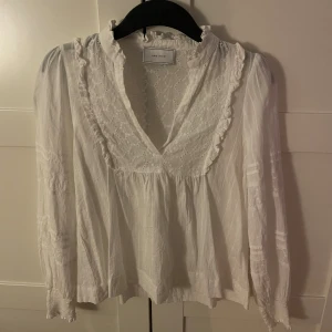 Vit blus med spets från Neo Noir - Romantisk vit blus från Neo Noir med broderade spetsdetaljer och volangkant vid v-ringningen. Blusen har långärmade ärmar med lätt puff och smock vid ärmslut. Tillverkad i tunn bomull med broderade mönster och en luftig passform.