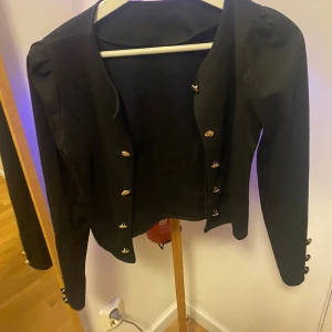 Svart kavaj med guldfärgade knappar - Stilren svart kavaj med snygga guldfärgade knappar framtill och vid ärmsluten. Jackan har en klassisk, rak passform och är långärmad. Perfekt för dig som gillar en clean look med lite extra detaljer. Materialet känns mjukt och har en lätt struktur.