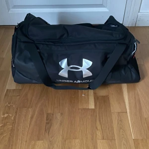 Svart träningsväska från Under Armour - Stor svart träningsväska från Under Armour med vitt logotryck på framsidan. Väskan har flera fack med dragkedja och justerbar axelrem. Tillverkad i slitstark polyester och rymmer allt du behöver till gymmet eller sportaktiviteter.