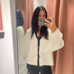 Vit stickad kofta med pärlemorknappar - Säljer en vit stickad kofta med pärlemorknappar och avslappnad passform. Koftan har långa ärmar och ribbade muddar vid ärmslut och nederkant. Perfekt att slänga över en topp för en chill look. Fråga gärna om fler bilder eller mått!