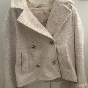 Vit teddyjacka från Nelly XS - Säljer en vit teddyjacka från Nelly i storlek XS. Jackan har dubbelknäppning med stora knappar, bred krage och två fickor framtill. Materialet är mjukt och fluffigt, perfekt för kyliga dagar. Snygg och trendig modell som ger en cool vibe.