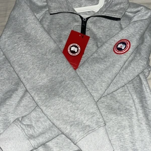 Grå half zip hoodie från Canada Goose - Snygg grå hoodie från Canada Goose med half zip och klassisk logopatch på bröstet. Bara testad hemma, aldrig använd helt ny! Pris kan diskuteras!
