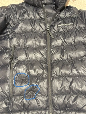 Svart Peak Performance dunjacka 140 - Svart dunjacka från Peak Performance med huva och broderad logga på bröstet. Jackan har två sidofickor med dragkedja, ribbade muddar och quiltad design. Perfekt för kalla dagar och har en snygg, sportig look. Stl 140, har två små defekter (se sista bild) därav priset