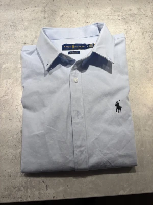 Ljusblå skjorta från Ralph Lauren - Snygg ljusblå skjorta från Ralph Lauren i klassisk passform. Skjortan har knappar framtill och den ikoniska broderade loggan på bröstet. Tillverkad i mjuk bomull, perfekt för en clean och stilren look. Krage med knappar och diskret design.