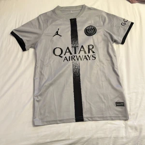 PSG fotbollströja Jordan x Qatar Airways - Snygg PSG fotbollströja i ljusgrått med svart vertikal rand och svarta detaljer. Jordan-logga och Paris Saint-Germain-emblem på bröstet, samt Qatar Airways tryck. Kortärmad och tillverkad i lätt, ventilerande polyester. Perfekt för dig som vill ha en clean och modern look på planen.