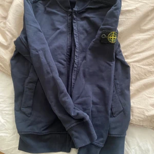  Stone Island junior collegetröja full zip - Snygg mörkblå full zip collegetröja från Stone Island med klassisk patch på ärmen. Jackan har dragkedja framtill, ribbade muddar och två fickor. Tillverkad i mjuk bomull som är skön att bära. Perfekt för dig som gillar streetwear och vill ha en clean look.