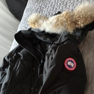Svart Canada Goose wyndham - Svart dunjacka från Canada Goose med fluffig beige päls på huvan och klassisk logopatch på ärmen. Jackan har lång ärm, dragkedja och tryckknappar framtill. Perfekt för kalla vinterdagar och har en snygg, tidlös look.köpt förra vintern 