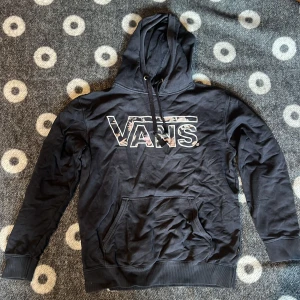 Svart hoodie från Vans  - Svart hoodie från Vans med stor logga i blommigt mönster på bröstet. Tröjan har huva med snörning och en klassisk magficka. Perfekt för dig som gillar streetwear och vill ha en chill stil.