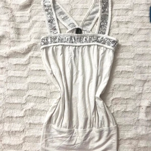 Vit kort klänning med paljettdetaljer - Säljer en vit kort klänning med breda axelband och snygga silvriga paljetter längs banden och upptill. Klänningen har en figurnära passform och är ärmlös, perfekt för dig som gillar att sticka ut med lite extra bling.