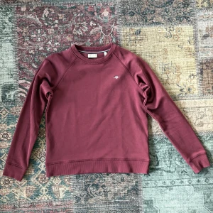 Vinröd sweatshirt från GANT - Snygg vinröd sweatshirt från GANT med diskret logga på bröstet. Tröjan har rund hals, ribbade muddar och är tillverkad i mjuk bomull. Perfekt för dig som gillar klassisk stil med en modern touch.
