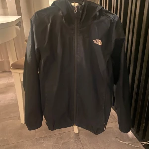 Svart vindjacka från The North Face - Svart vindjacka från The North Face med huva och hel dragkedja framtill. Jackan har en stilren design med logga på bröstet och elastiska muddar vid ärmslut. Perfekt för blåsiga dagar och enkel att matcha med din streetwear. Inga tecken på slitage eller liknande förutom att detaljen på dragkedjan har gått av 