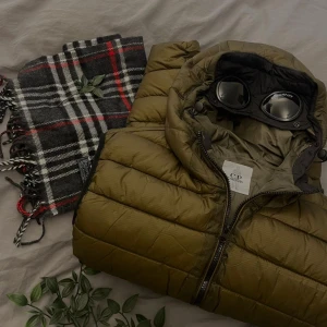 Olivgrön pufferväst från C.P. Company - Säljer en olivgrön pufferväst från C.P. Company med ikonisk goggle-hood. Jackan har dragkedja framtill och är tillverkad i polyester och nylon. Perfekt för dig som gillar streetwear och vill sticka ut med en unik detalj på huvan.