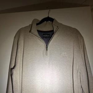 Beige stickad half zip från Jean Paul - Snygg beige stickad tröja från Jean Paul med halv dragkedja och broderad logga på bröstet. Tröjan har en hög krage och långa ärmar, perfekt för dig som gillar stilren och enkel design. Passar dig som vill ha en chill och klassisk look. 