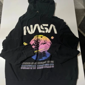 Svart NASA hoodie med färggrafik - Svart hoodie från DIVIDED med stor NASA-logga och färgglad grafik på bröstet. Trycket har inslag av lila, rosa, blått och gult, samt texten 'Scientific Discoveries'. Tröjan har huva med snörning och långa ärmar. Perfekt för dig som gillar rymden och coola prints.