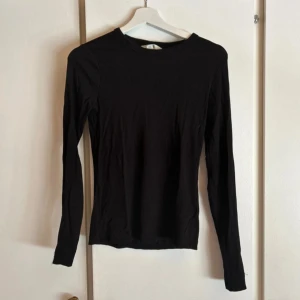 Svart långärmad tröja i bomull - En enkel svart långärmad tröja med rund hals. Modellen är slim och sitter snyggt mot kroppen. Tillverkad i mjuk bomull som är skön att ha på sig. Perfekt att styla med jeans eller under en jacka. Passar dig som gillar minimalistisk och clean stil.