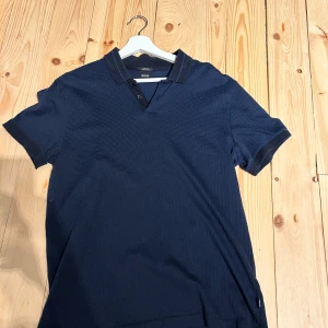 Marinblå pikétröja från BOSS - Snygg marinblå pikétröja från BOSS i slim fit med diskret textur och klassisk krage med två knappar. Tillverkad i mjuk pima bomull som känns skön mot huden. Kragen har en tunn blå linje som detalj och korta ärmar med ribbade muddar. 