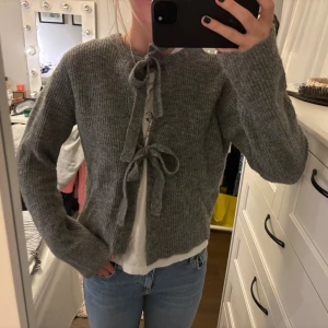 Grå stickad kofta från Object - Supermysig grå stickad kofta från Object i storlek S. Koftan har ribbad struktur, långa ärmar och två snygga knytband framtill. Perfekt att slänga över en t-shirt för en chill look. Passar dig som gillar enkel och stilren design.