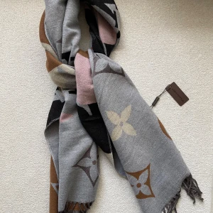 Vuitton grå halsduk med logomönster - Lyxig och stor halsduk från Vuitton i grått med ikoniska blommönster och -logga. Färgerna går i grå, brun, beige, svart och ljusrosa toner. Materialet är mjukt och stickat med snygga fransar i kanterna. Perfekt att svepa runt axlarna för en trendig look.