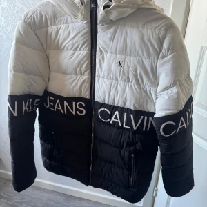 Svartvit pufferjacka Calvin Klein Jeans - Snygg pufferjacka från Calvin Klein Jeans i svart och vitt med stor logotyp längs bröst och ärmar. Jackan har huva, dragkedja framtill och två fickor med dragkedja. Perfekt för dig som gillar streetstyle och vill sticka ut med en fet logga. Storlek: 14 år. Produkten finns på flera sidor.