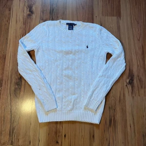 Vit kabelstickad tröja Ralph Lauren - Snygg vit kabelstickad tröja från Ralph Lauren i 100% bomull. Klassisk rund hals och långärmad modell med marinblå broderad logga på bröstet. Perfekt för dig som gillar stilrena och tidlösa plagg med preppy vibe. Passar M/175. Finns en liten fläck på ärmen som går att tvätta 🧽 