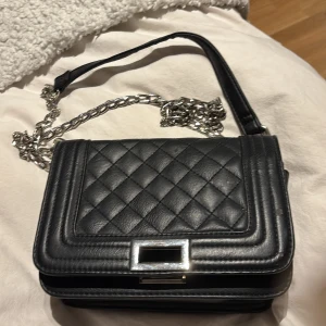 Svart väska med kedja - Snygg svart väska i skinn med silverfärgad kedjerem och silverspänne. finns skavanker på den då den är använd. se bilder.