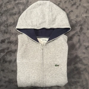 Lacoste zip hoodie - Grå - Snygg grå hoodie från Lacoste Sport med hel dragkedja och klassisk krokodillogga broderad på bröstet. Tröjan har huva med mörkblå insida och långa ärmar. Perfekt för chill dagar och sportiga outfits. Storlek S
