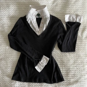 Ralph Lauren tröja - Svart v-ringad tröja från Lauren Ralph Lauren med insydd vit skjortkrage och manschetter. Tröjan har lång ärm och en slim passform som sitter snyggt. Materialet är mjuk bomull och skjortdetaljerna är guldiga.