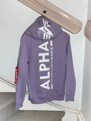 Lila hoodie från Alpha Industries - Snygg lila hoodie från Alpha Industries med stor vit logga på ryggen och mindre logga fram. Klassisk huva med dragsko, magficka och en unik ficka med röd tagg på ärmen. Perfekt för dig som vill sticka ut med streetwear-vibe. Typ aldrig använd