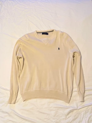 Beige tröja från Polo Ralph Lauren - Klassisk beige tröja från Polo Ralph Lauren med rund halsringning och den ikoniska svarta logotypen broderad på bröstet. Tröjan har långa ärmar och ribbade muddar vid ärmslut och nederkant. Perfekt för en stilren och avslappnad look.