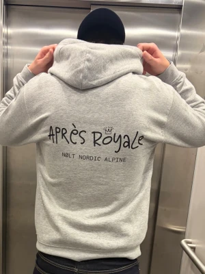 Grå hoodie Après Royale - Snygg grå hoodie från Nølt Nordic Alpine med stor svart text 'Après Royale' och logga på ryggen. Klassisk passform med huva och mjukt material, perfekt för en avslappnad stil. Stora tryckdetaljer ger en cool vibe.