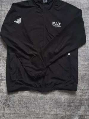 Svart vindjacka från EA7 Armani - Snygg svart vindjacka från EA7 Emporio Armani med dragkedja framtill och vita logotryck på bröstet. Jackan har elastiska muddar vid ärmslut och nederkant samt två sidofickor med dragkedja. Perfekt för en sportig och stilren look. FINNS LUVA som inte syns i bilden