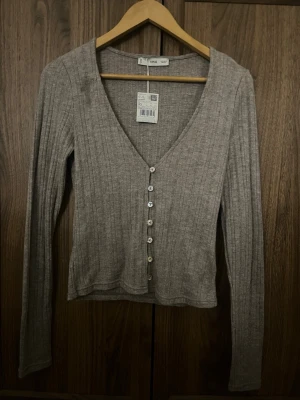 Ribbad beige kofta från MANGO - Säljer en ribbad grå kofta från MANGO med v-ringning och pärlemorknappar framtill. Koftan har långa ärmar och är tillverkad i ett mjukt, stretchigt material som sitter snyggt på kroppen. Perfekt att styla med jeans eller kjol.