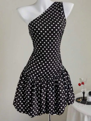 Svart prickig oneshoulder klänning - Säljer en svart kortklänning med vita prickar och oneshoulder-design. Klänningen har en figurnära överdel och en volangig kjol som ger en lekfull look. Perfekt för dig som vill sticka ut med ett klassiskt men trendigt mönster. Använd en gång🖤🤍