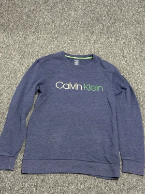 Blå sweatshirt från Calvin Klein - Mysig blå sweatshirt från Calvin Klein med logga i vitt och grönt på bröstet. Tröjan har rund halsringning, långa ärmar och ribbade muddar. Perfekt för en avslappnad och stilren look.