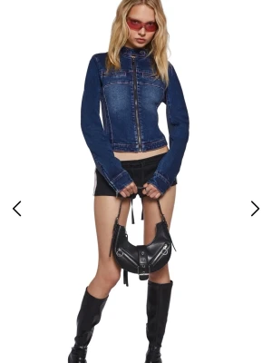 Mörkblå jeansjacka från Sugar Thrillz - Slutsåld Snygg mörkblå jeansjacka från Sugar Thrillz med rosa sömmar, hög krage och dragkedja framtill. Jackan har en figurnära passform och coola detaljer som bröstficka med dragkedja. Perfekt för en trendig streetstyle-look. Justerbar baktill och nedtill vid ärmarna. Oanvänd! Säljer då jag redan har en i xxs! Denna passar definitivt XS och kanske även S om man vill ha extra tight! 