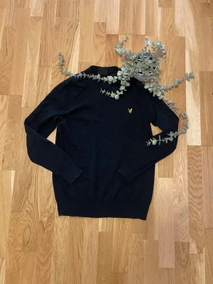 Mörkblå stickad tröja Lyle & Scott - Mörkblå stickad tröja från Lyle & Scott med klassisk gul broderad logotyp på bröstet. Tröjan har rund halsringning, långa ärmar och ribbade muddar vid ärmslut och nederkant. Perfekt för dig som gillar stilrena och tidlösa plagg.