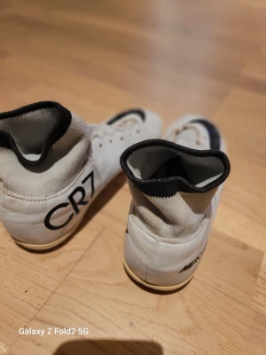 Nike CR7 vita fotbollsskor - Säljer ett par vita Nike Mercurial fotbollsskor med CR7-tryck på hälen och stora svarta swoosh-loggor på ovansidan. Skorna har högt skaft i stickat material och svart kant upptill. Perfekta för dig som vill sticka ut på planen.