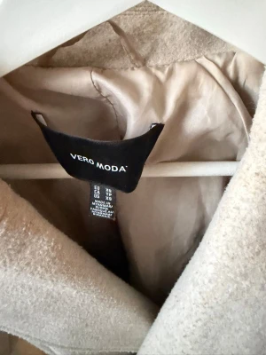 Beige kappa med knytskärp Vero Moda - Stilren beige kappa från Vero Moda med klassisk krage och knytskärp i midjan. Kappan har en mjuk yta och är helfodrad för extra komfort. Perfekt för dig som vill ha en enkel men snygg look till hösten eller våren.