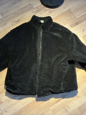 Svart teddyjacka från Weekday - Mysig svart teddyjacka från Weekday med dragkedja framtill och hög krage. Jackan har en fluffig och mjuk yta i teddy-material och två sidofickor. Perfekt för kalla dagar när du vill vara både varm och snygg.